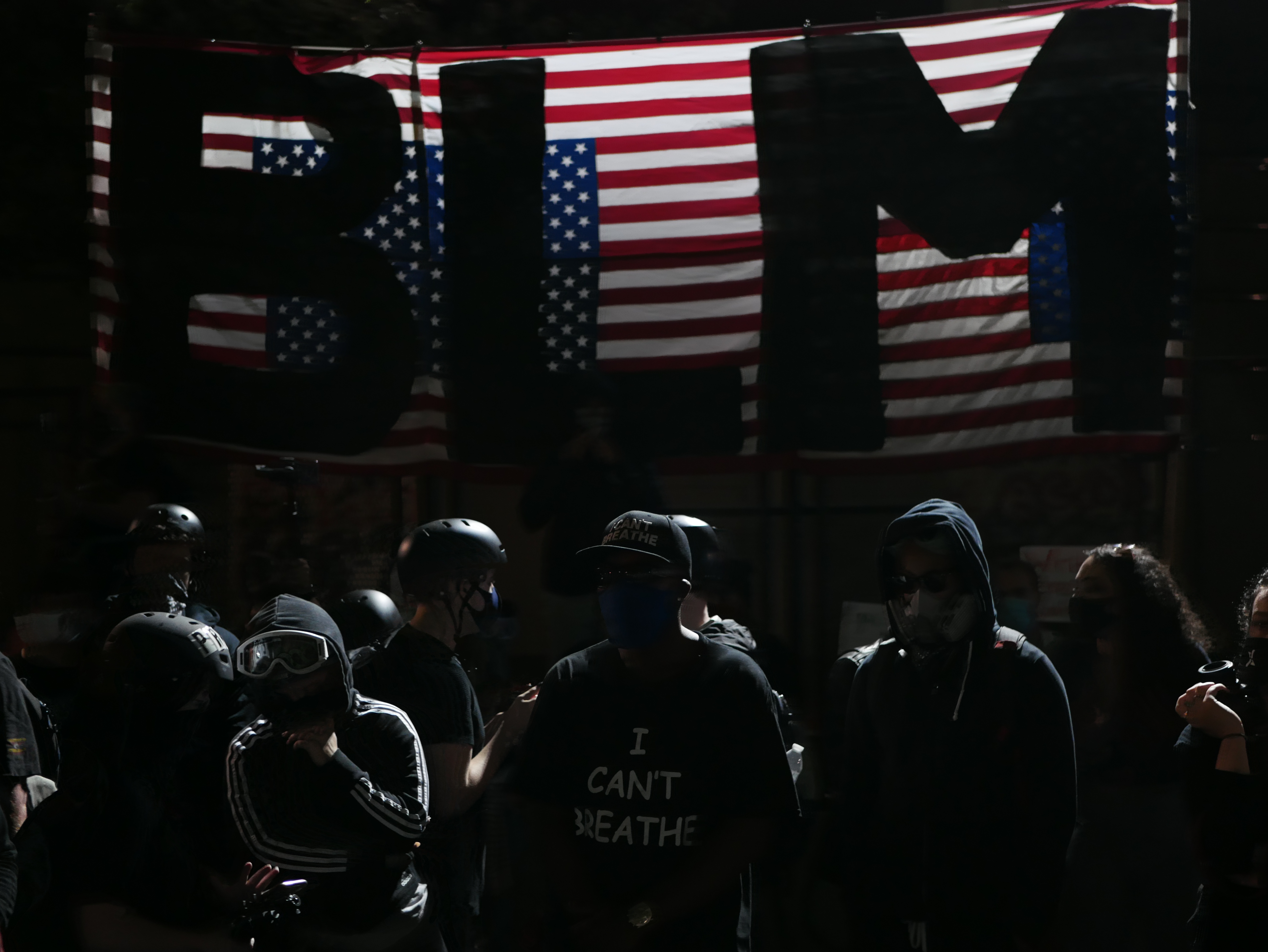 Photograph of protesters in front of an American flag with BLM in black letters written on it.