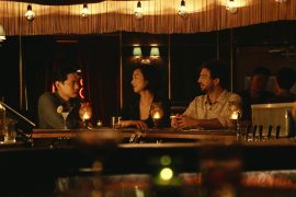 photo of film scene. Three people are at a bar. Two are facing each other and the other is facing the other two, but in the conversation.
