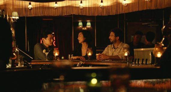 photo of film scene. Three people are at a bar. Two are facing each other and the other is facing the other two, but in the conversation.